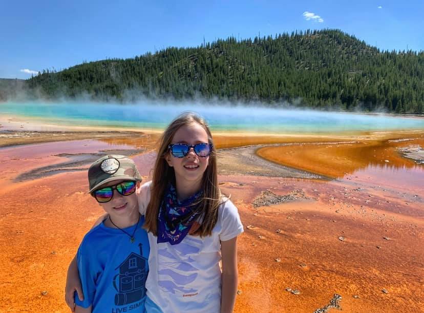 Two kids stand smiling with the Grand Pristmatic behind them at Yellowstone National Park.