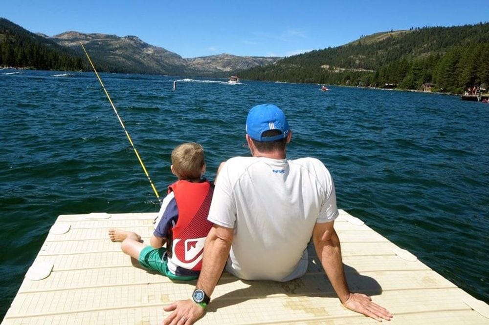 A boy holding a fishing pole sits near his dad on a dock.