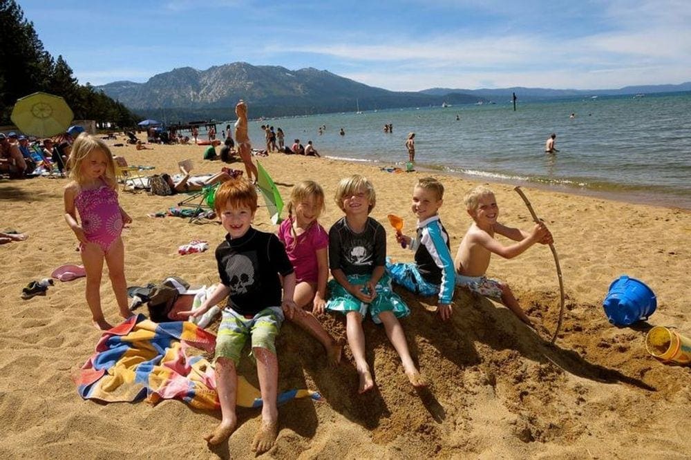 Six kids sit together, smiling, while enjoying a sunny day at the beach along Lake Tahoe, with mountains in the distance.