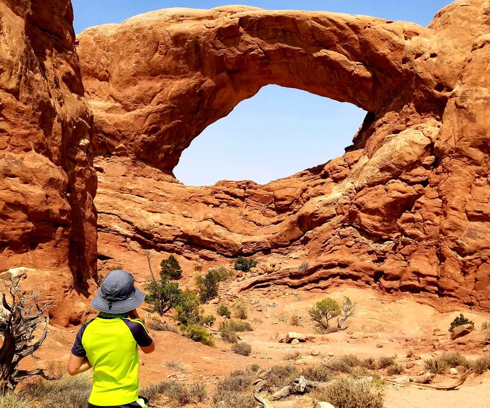 A young boy wearing a yellow shirt and a blue sun hat looks at the iconic Utah arches at Arches National Park, one of the best things to do in Moab with kids.