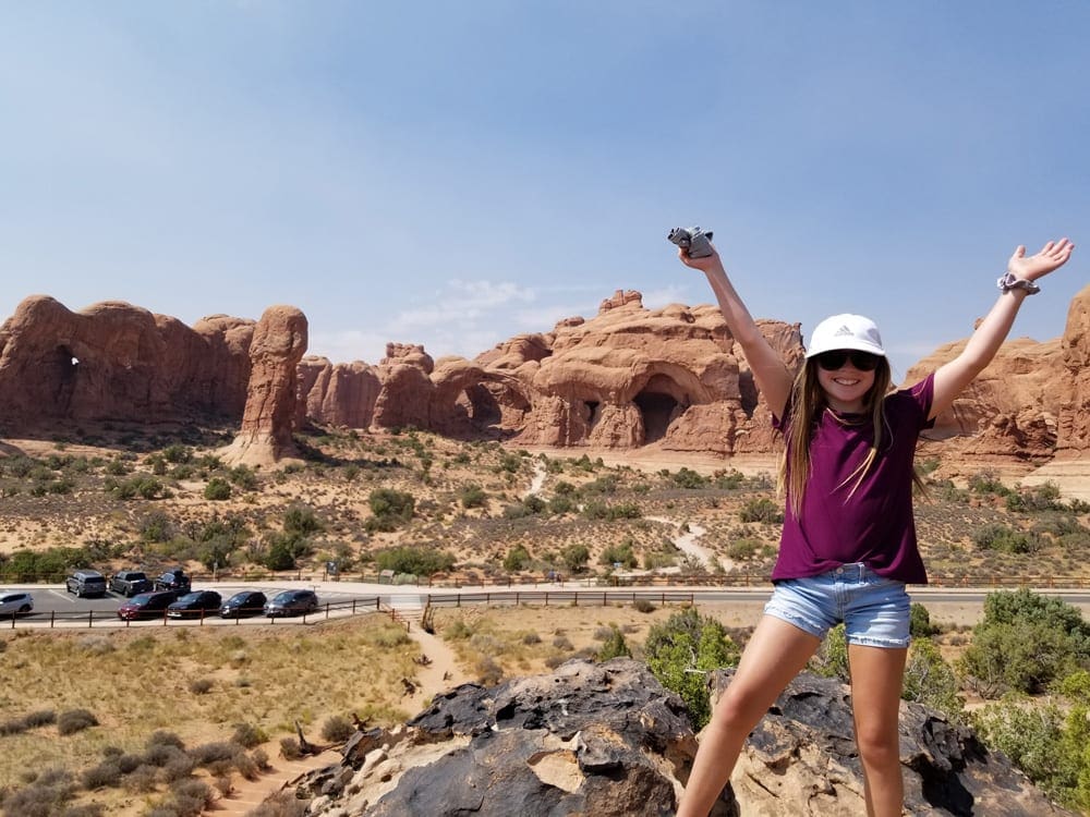 A young girls excitedly throws her hands in the air with the iconic Utah red rock formations in the distance while exploring Moab, one of the best vacation spots in the US to impress teens and tweens.