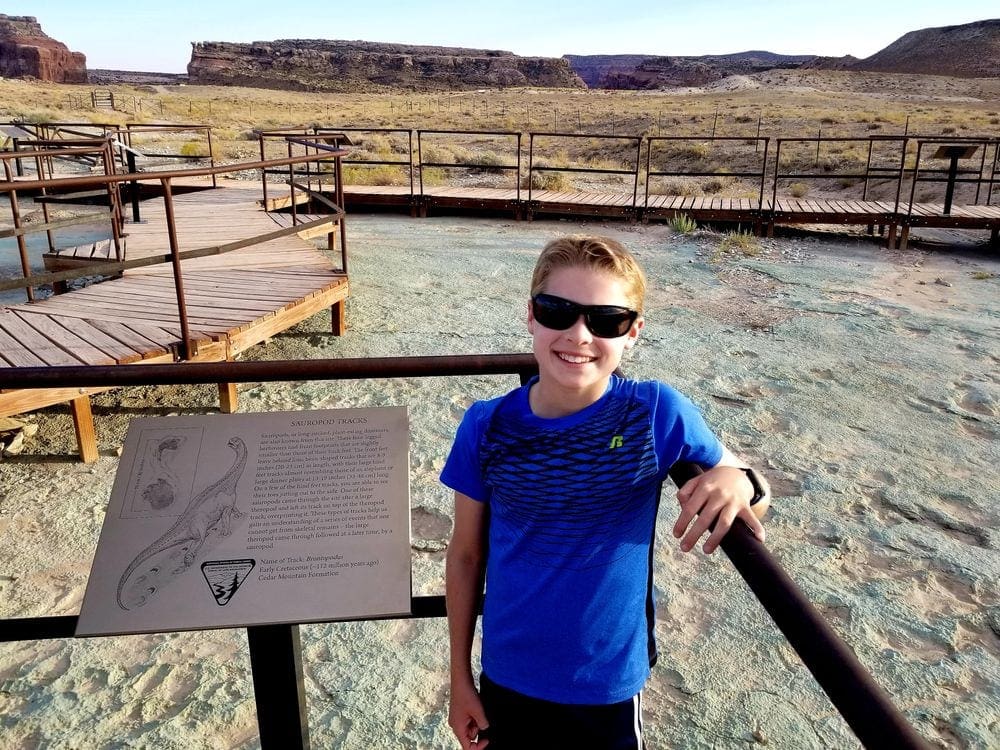 A young boy wearing a blue shirt and sunglasses stands near a sign featuring a dinosaur, while wandering the Dinosaur Trail in Utah, one of the best affordable summer vacations in the United States with kids.
