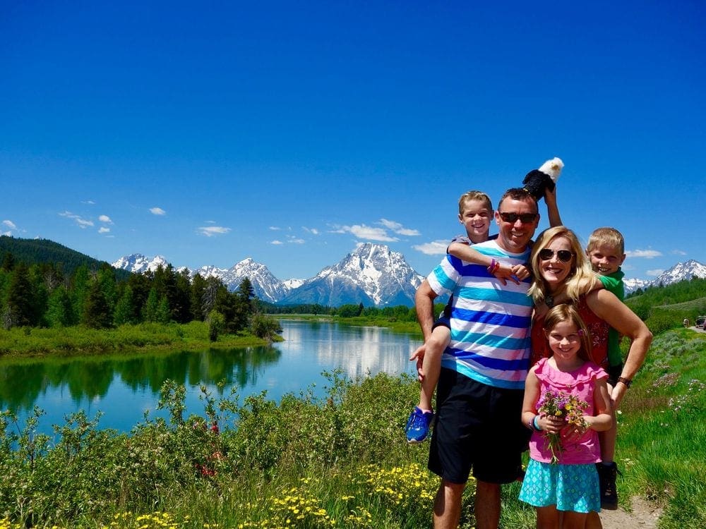 A family of five stands together smiling on a beautiful sunny day near Jenny Lake with the Grand Tetons mountains in the distance.