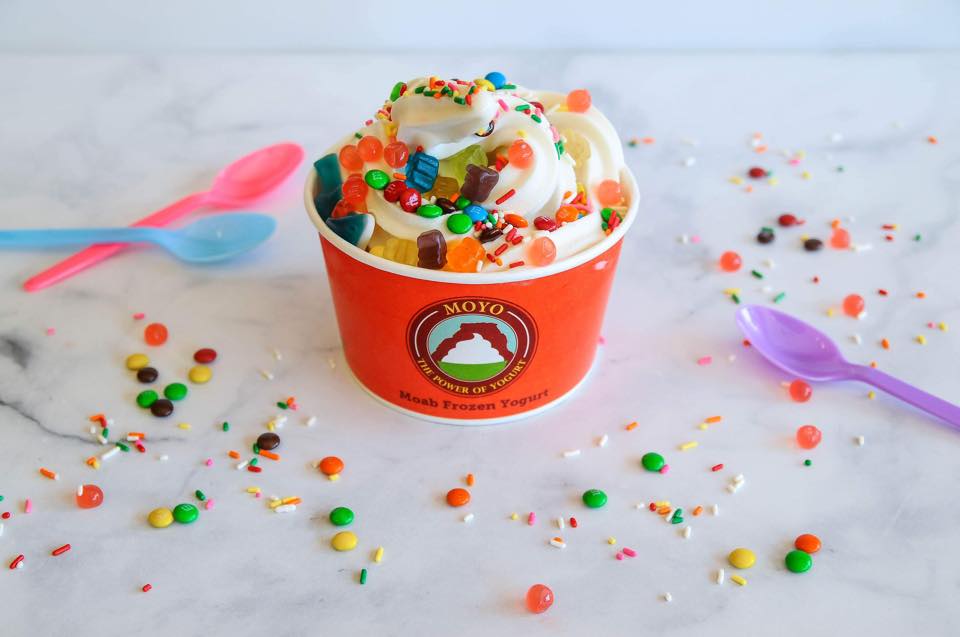 A cup of frozen yogurt with sprinkles sits on a white table surounded by more sprikles and colorful spoons.