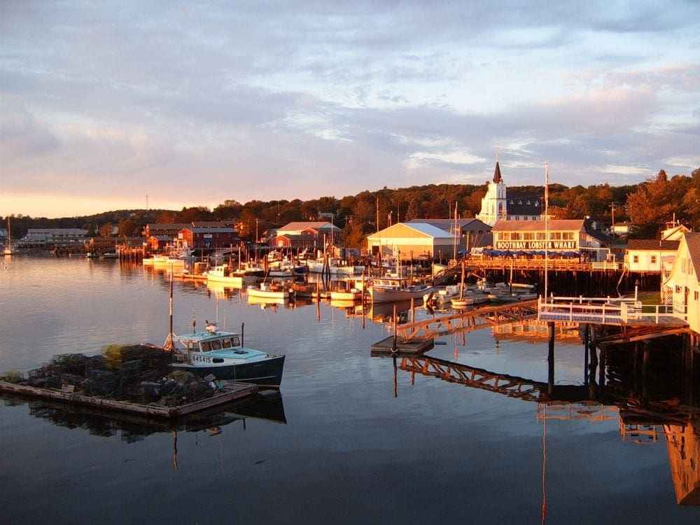 The town of Boothbay Harbor shines at dusk, with charming buildings and a bay holding several boats.
