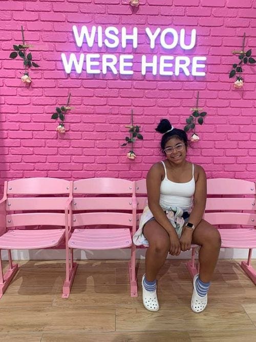 A young girl sits in a pink chair in front of a pink wall with a neon pink sign reading "Wish You Were Here".