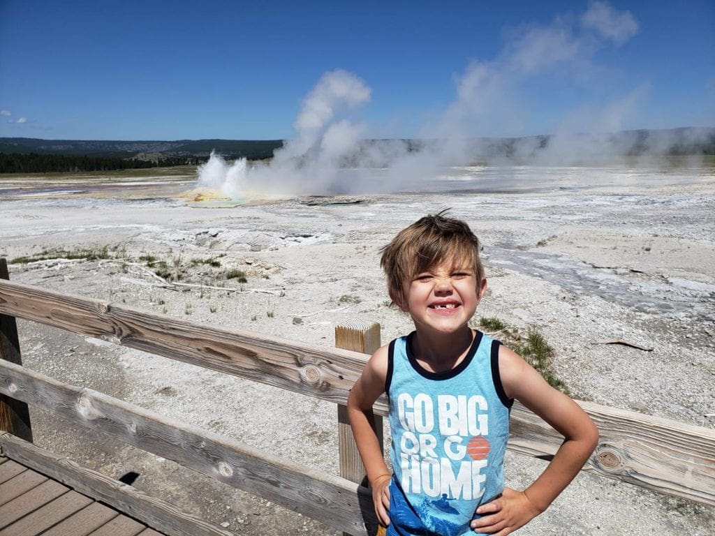 A young boy poses with a huge smile in front of a geyser in Yellowstone National Park.