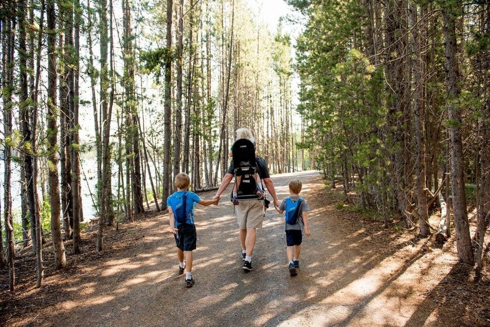 A dad, wearing one of his kids on his back, holds the hands of two other children on either side of him, while hiking, one of the best things to do in Grand Tetons National Park with kids.
