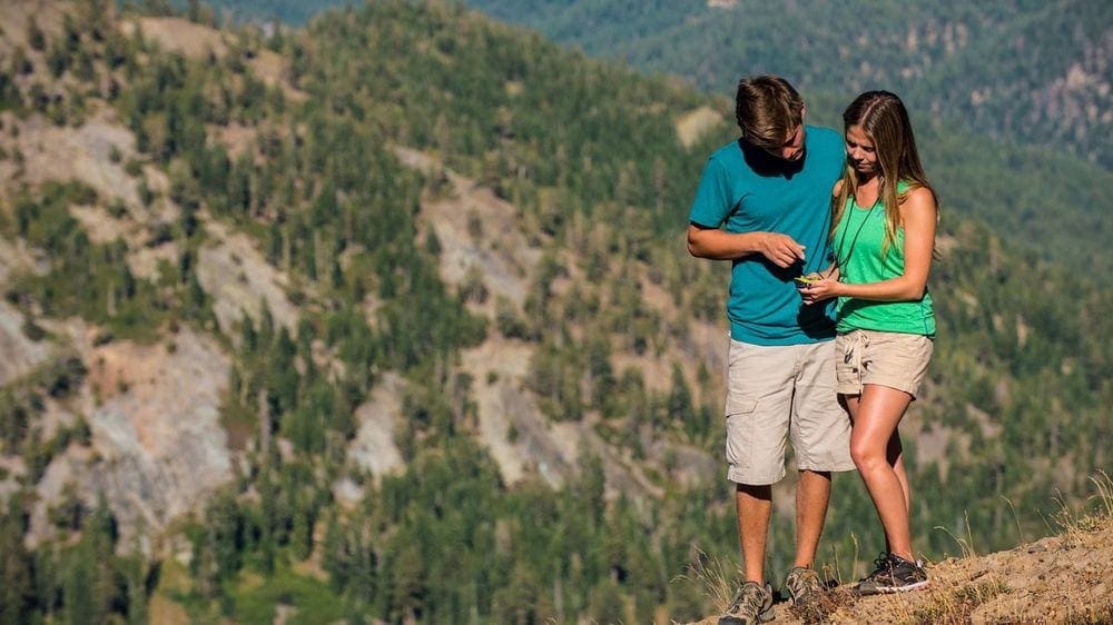 Two teens look at a device while exploring a mountain terraine in search of a geocache.