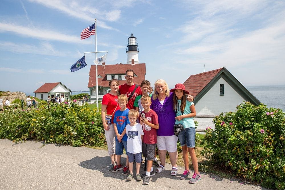 A family of eight stands together smiling in front of a lighthouse in Cape Elizabeth, Maine.