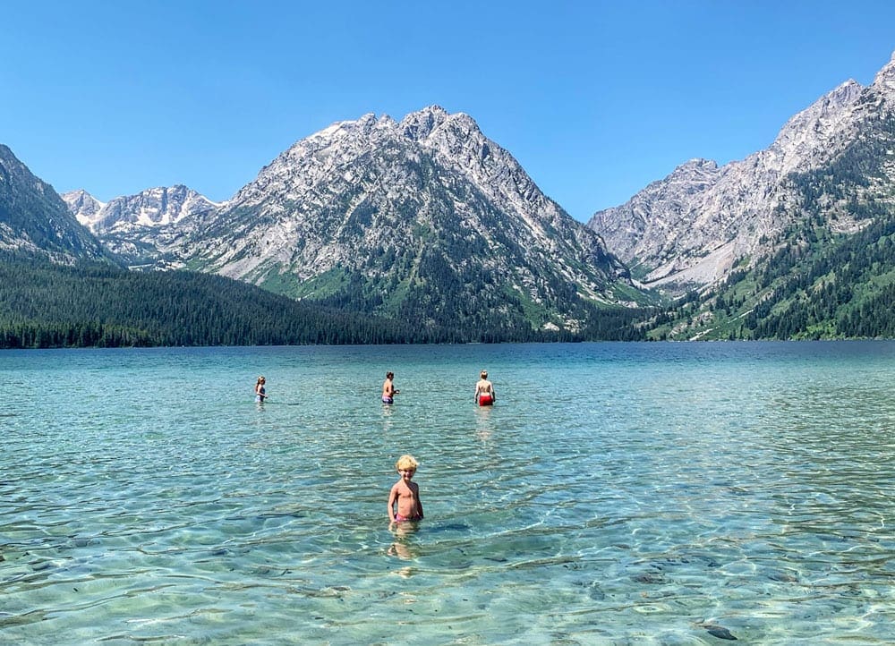 Several kids play in a shallow, clear blue lake with snow-capped mountains from the Grand Teton range behind them.