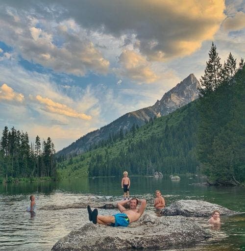 Four kids swim while their dad rests on a large boulder in String Lake, one of the best things to do in Grand Tetons National Park with kids.
