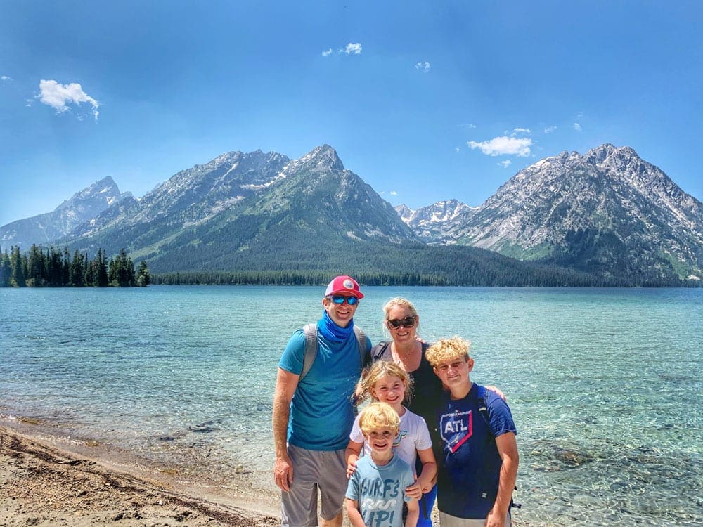 A family of four poses together along a lake with the Grand Tetons behind them.