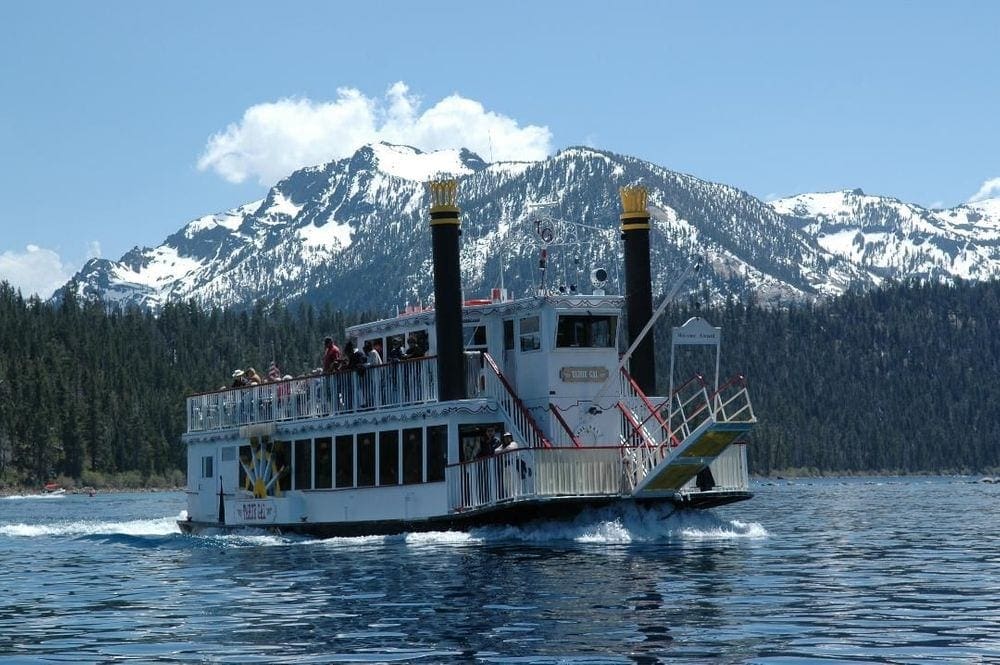 A large boat, called the Tahoe Gal, carries passangers on a cruise while exploring the waters of Lake Tahoe with snowcapped mountains in the distance.