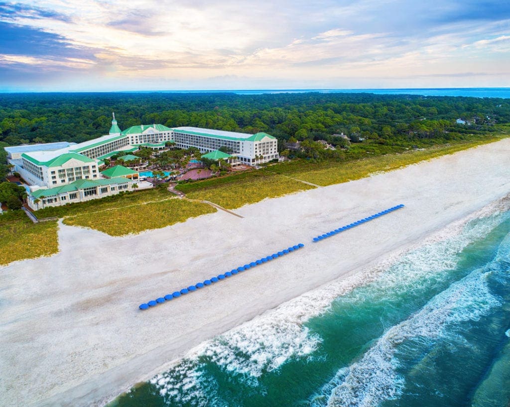 An aerial view of the oceanside The Westin Hilton Head Island Resort & Spa, featuring its iconic green roof and a row of blue beach umbrellas along the beach.