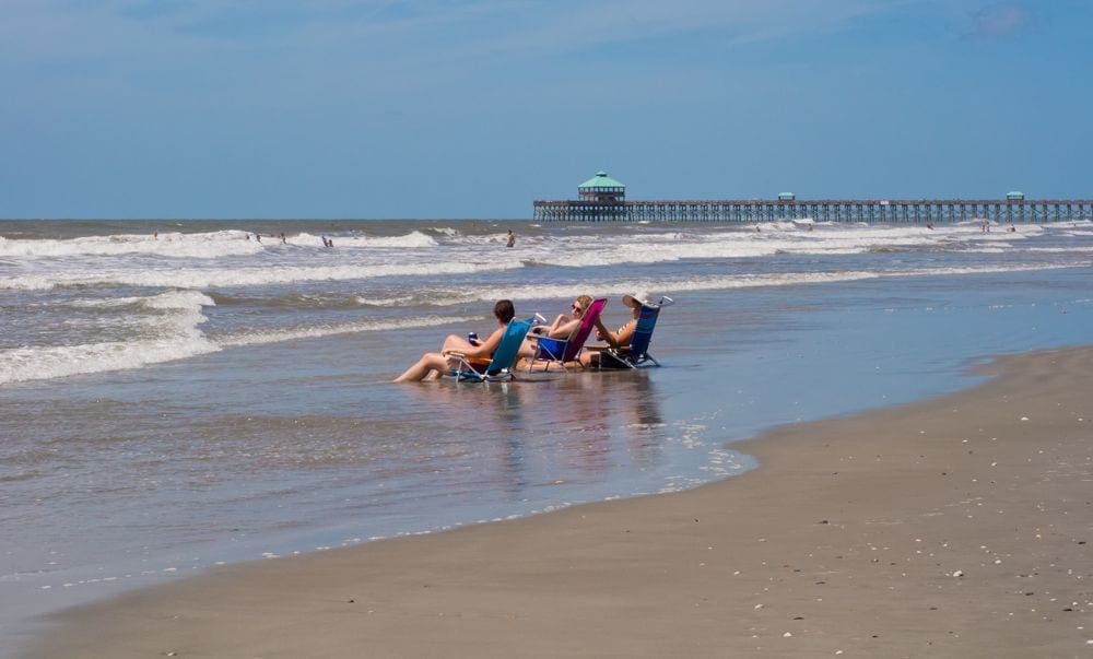 Three adults sit in beach chairs along the shores of Folly Island with the elevated boardwalk in the distance.