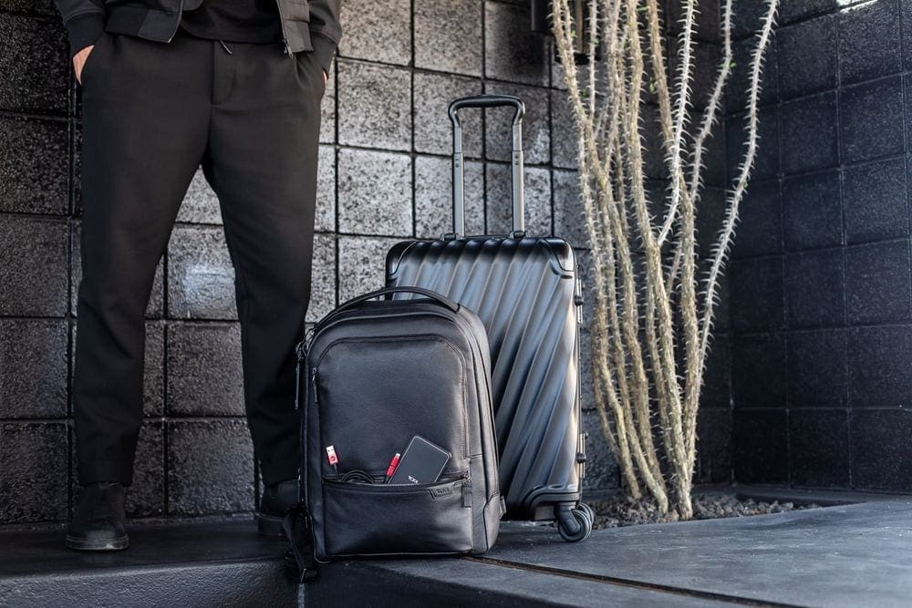 On the left, a pair of men's legs stand beside a black Tumi backpack and a black Tumi carry-on.