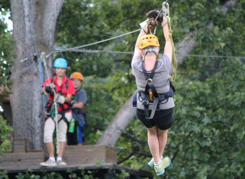 A person wearing a yellow helmet ziplines towards two invidiuals waiting on the treetop platform.