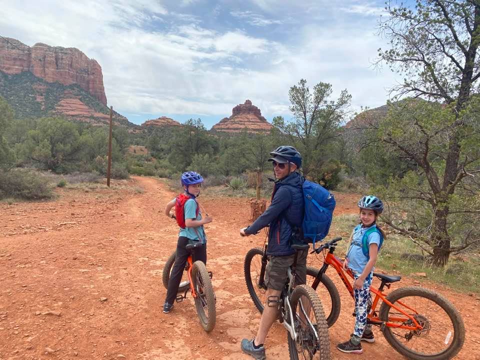 A dad and two kids perched on bikes look behind them at the camera while the iconic Sedona red rocks in the distance, a must see on any Sedona itinerary for families.