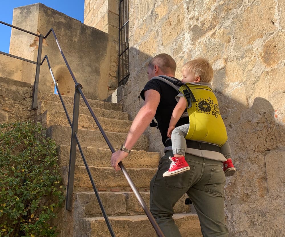 A dad carrying his son on his back in a yellow backpack walks up a flight of outdoor stairs in Portugal.