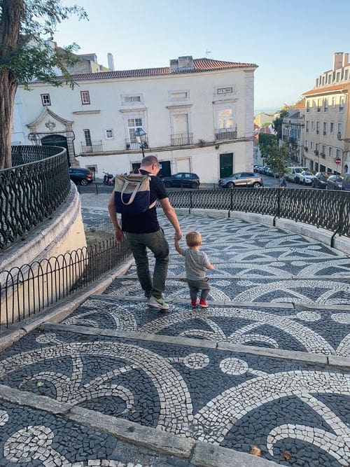 A dad holds the hand of his young son while walking down a blue and white tiled street in Lisbon.