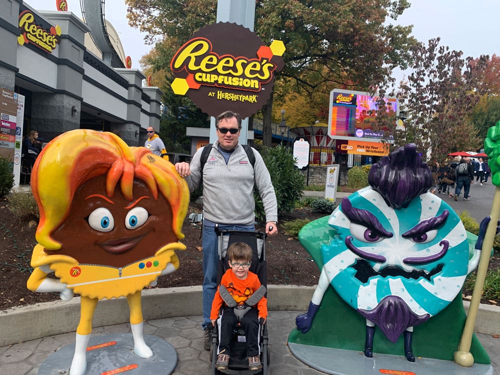 A dad stands behind his son, sitting in a stroller, between two mascots at Hershey Park, one of the best Memorial Day Weekend getaways from Washington DC for families.