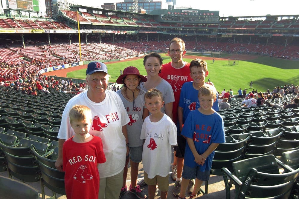 A family of eight poses together with Fenway Park in the background as they enjoy a baseball game on a sunny day in Boston, one of the best vacation spots in the US to impress teens and tweens.