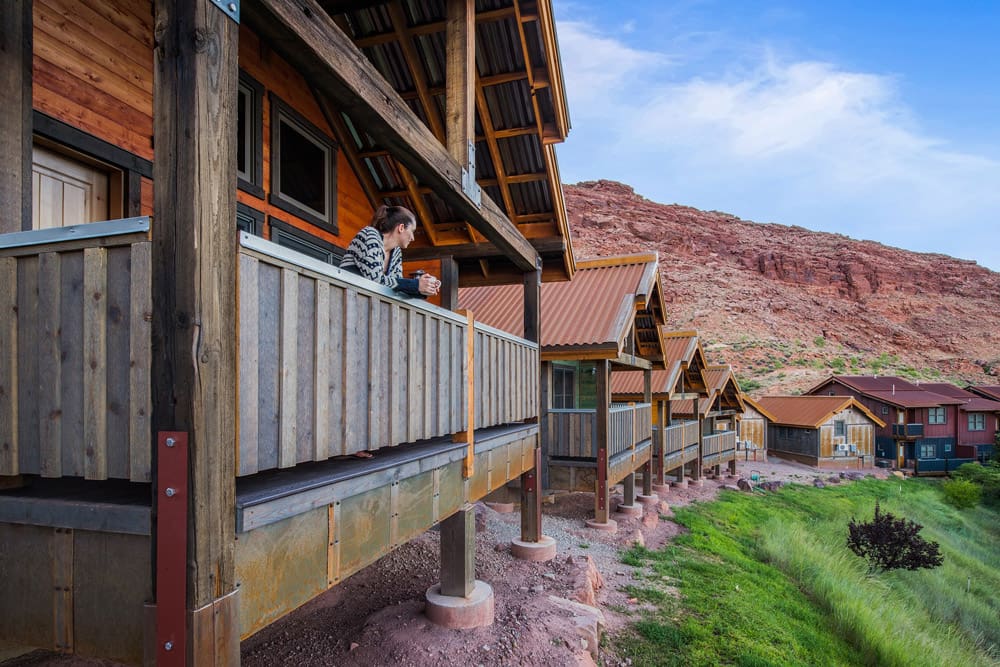 A woman hold a cup of coffee leans over a balcony to enjoy the view at the Moab Springs Ranch, one of the options for hotels in Moab for families.