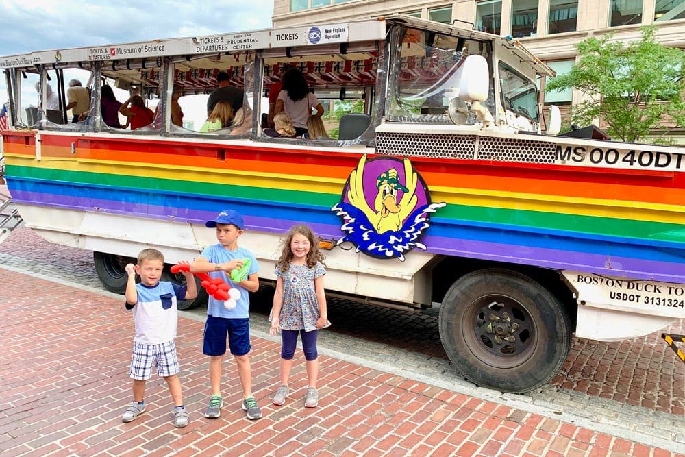 Three kids stand ashore in front of the Boston Duck Tour boat in Boston, one of the best Fourth of July destinations for a family trip.