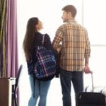 A woman wearing a backpack looks at a man pulling a suitcase in a hotel room on their romantic getaway.