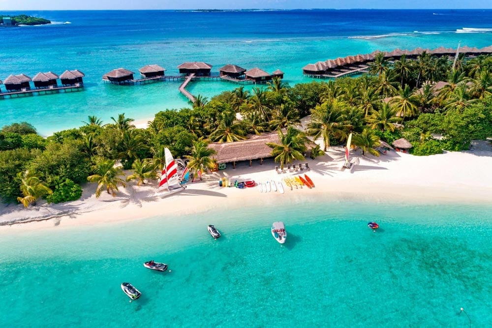 An aerial view of the Sheraton Maldives Full Moon Resort, one of the best family hotels in the Maldvies, featuring a pristine beach, several water crafts, and lush palms.