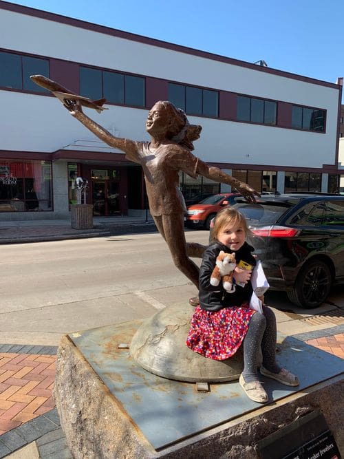 A young girl snuggles a stuffed animal cat while sitting near a statue of a girl flying plane in Eau Claire, Wisconsin.