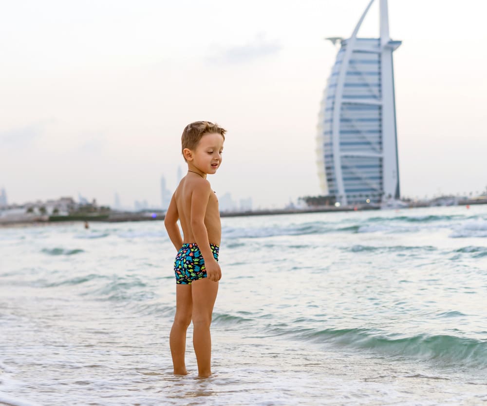 A young boy wearin swim trunks stands on a beach with one of the Best Family Hotels in Dubai in the distance.