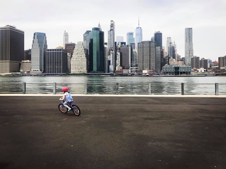 A young girl rides her bike with the Manhattan skyline behind her.