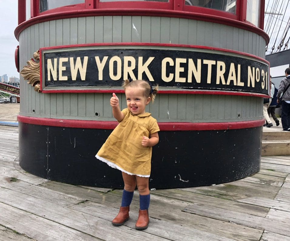 A young girl in a yellow dress gives a thumbs up outside the New York Central harbor, one of the best stops on our NYC itinerary with kids.