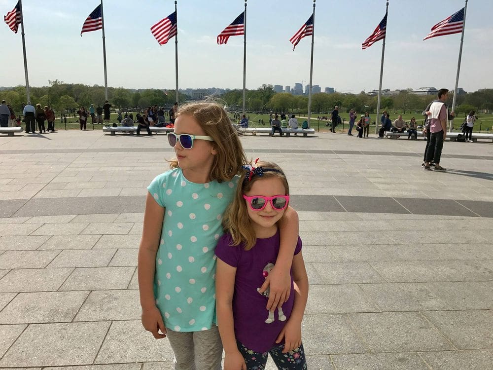 Two girls, wearing sun glasses, stand together with several American flags in the background.