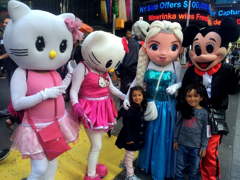 Two kids stand with characters such as Hello Kitty, Elsa, and Mickey Mouse while exploring Times Square, one of the best things to do New York City with young kids.