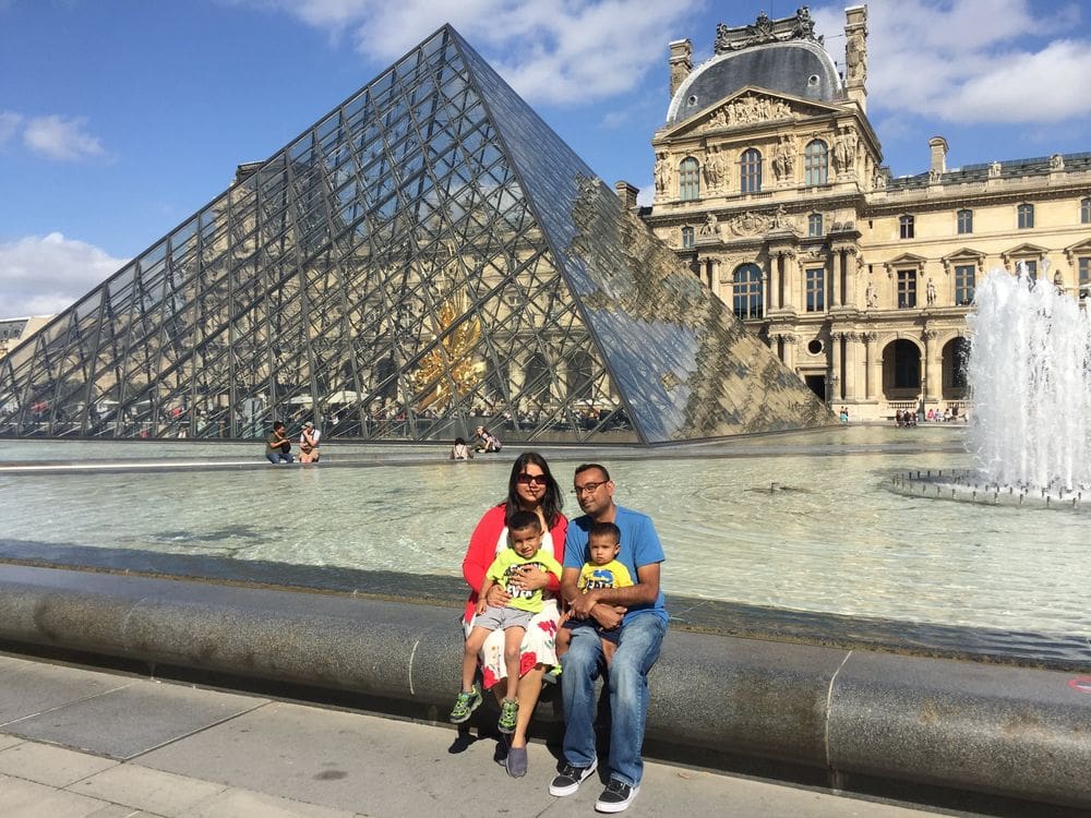 A family of four sits together on the edge of the fountain with the iconic Lourve pyramid behind them. 