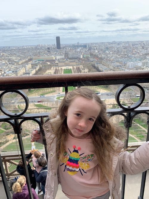 A young girl poses at the top of the Eiffel Tower with a grand view of Paris behind her.