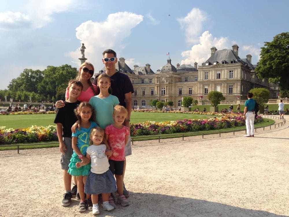 A family of six poses together with the Palace of Versailles behind them in France, one of the top places in travel in 2023 with kids.