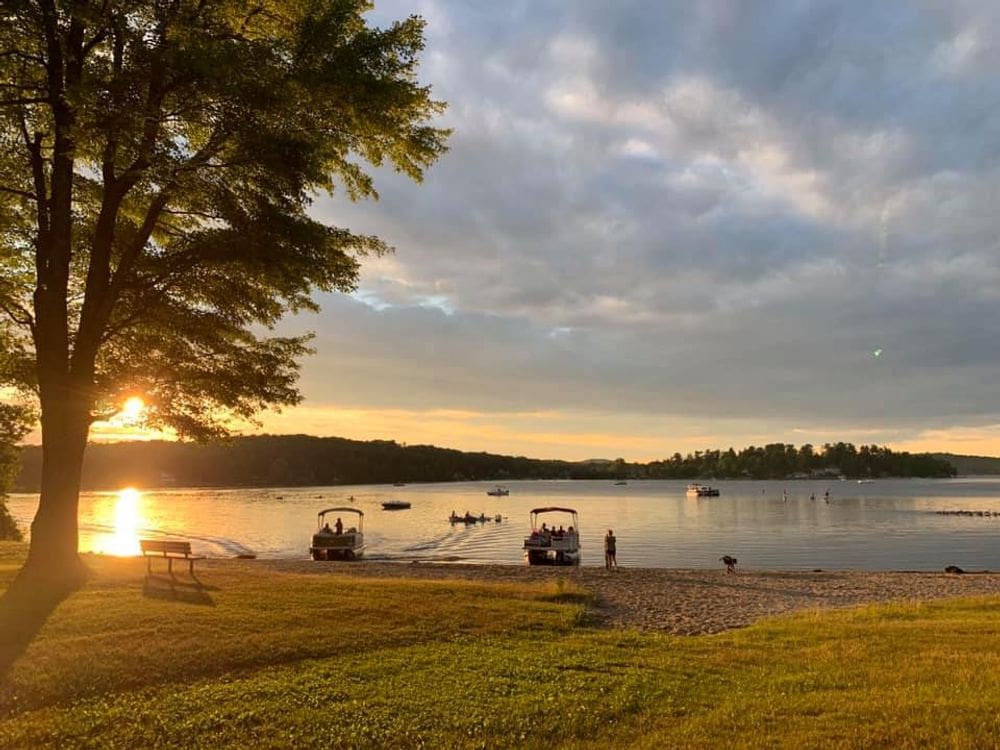 Several pontoons and swimmers enjoy a sunset at Morris Beach on Bantam Lake, one of the best lakes for a family vacation within 4 hours of NYC.