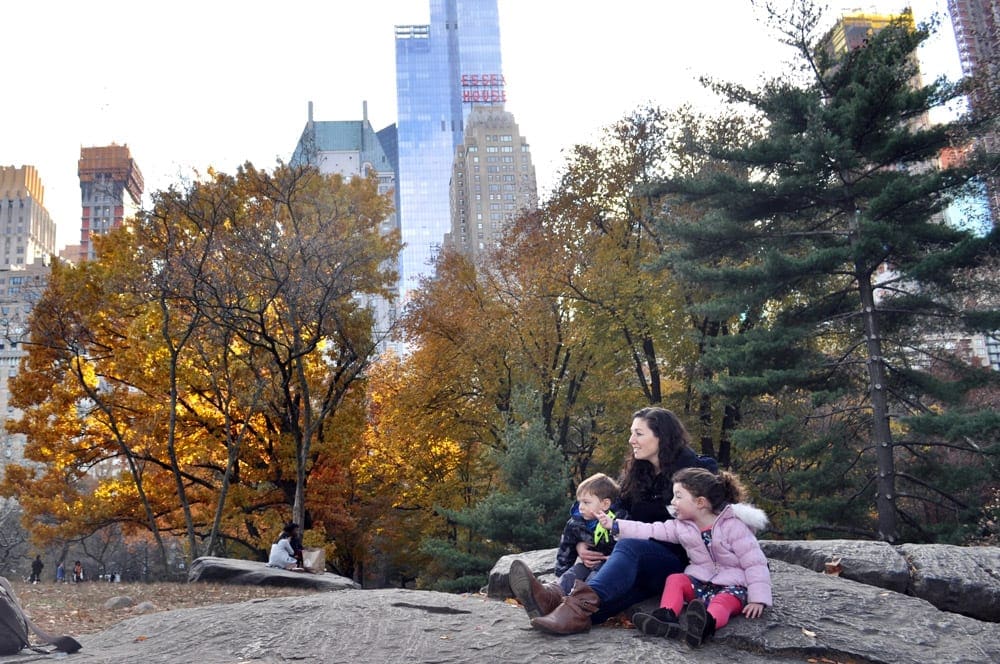 A mom and two kids sit on a large rock surrounded by fall foliage while exploring Central Park, one of the best things to do New York City with young kids.