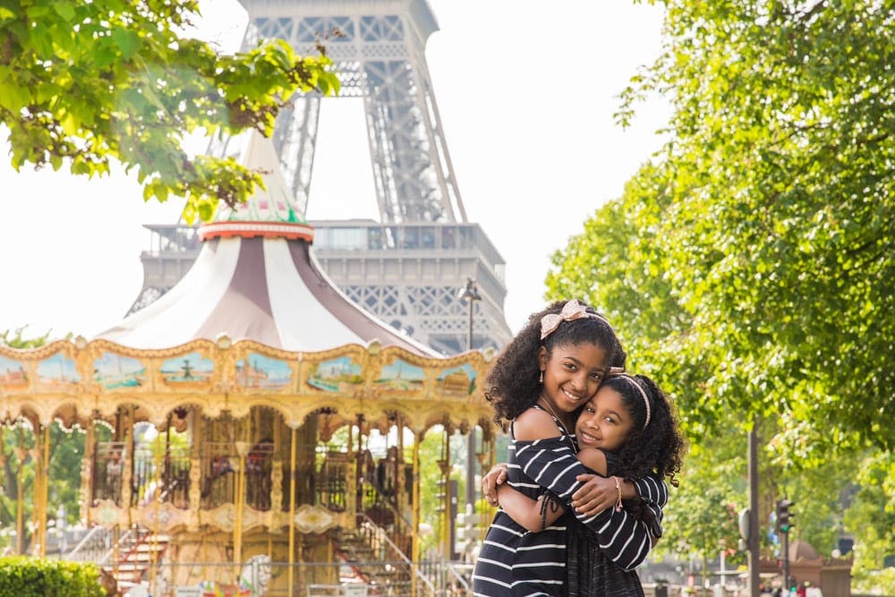 Two sisters of color embrace with Paris' iconic carousel and Eiffel tower behind them, one of the top places in travel in 2023 with kids.