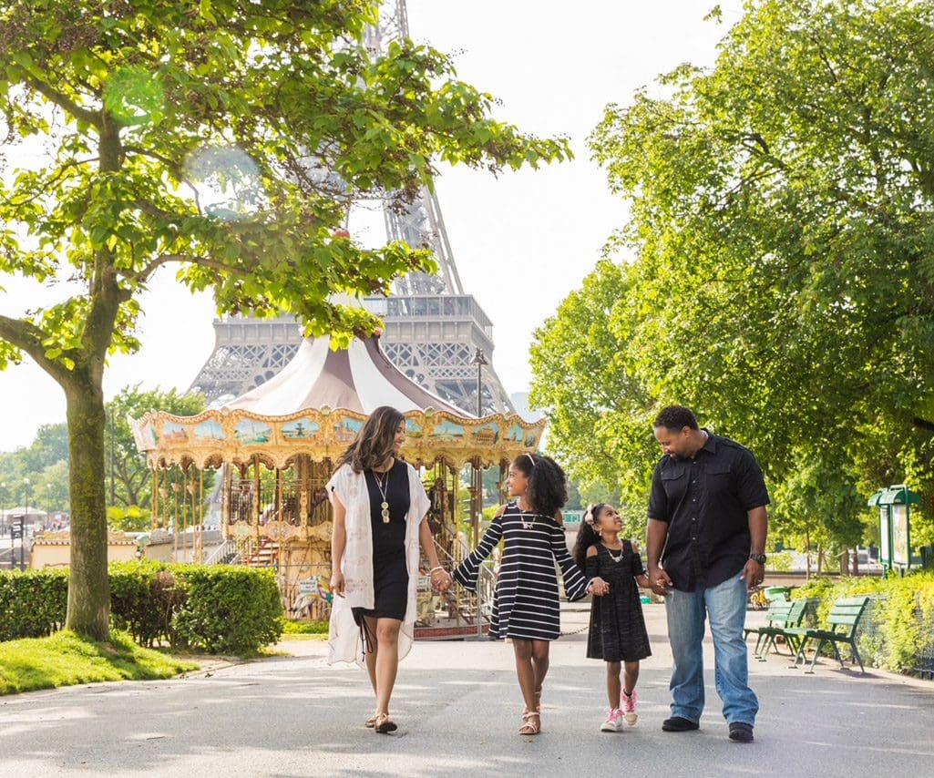 A family of four walks hand -in-hand with the iconic Paris carousel and Eiffel Tower in the distance.