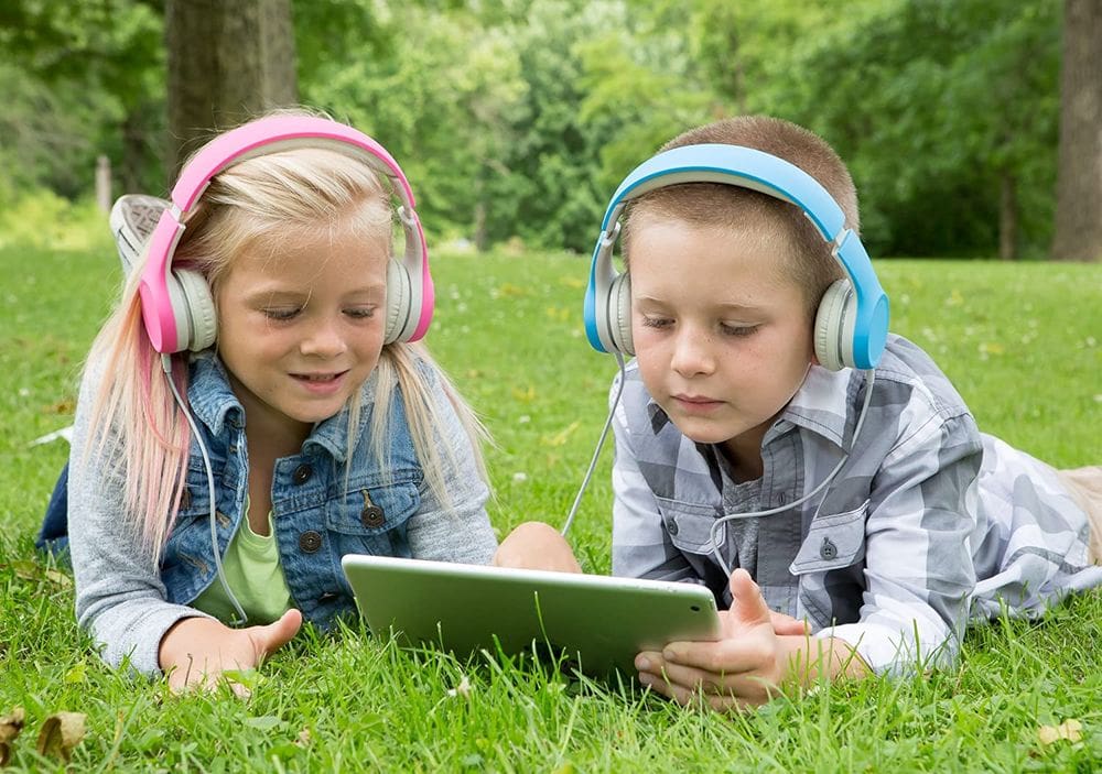 A girl and a boy both sport the Snug + Play Kids Headphones, one in blue and one in pink, while watching something on a tablet.