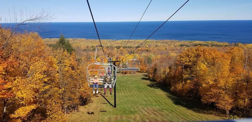 Two people ride on a chair lift up the Porkies with a view of brilliant fall colors and Lake Michigan behind them, one of the best places to see fall colors in the US for families.
