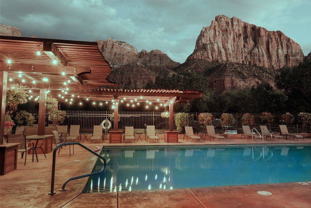 The outdoor pool, with expansive pool deck and loungers at Cable Mountain Lodge. Resort buildings behind the pool, as well as massive desert rock formations in the distance.