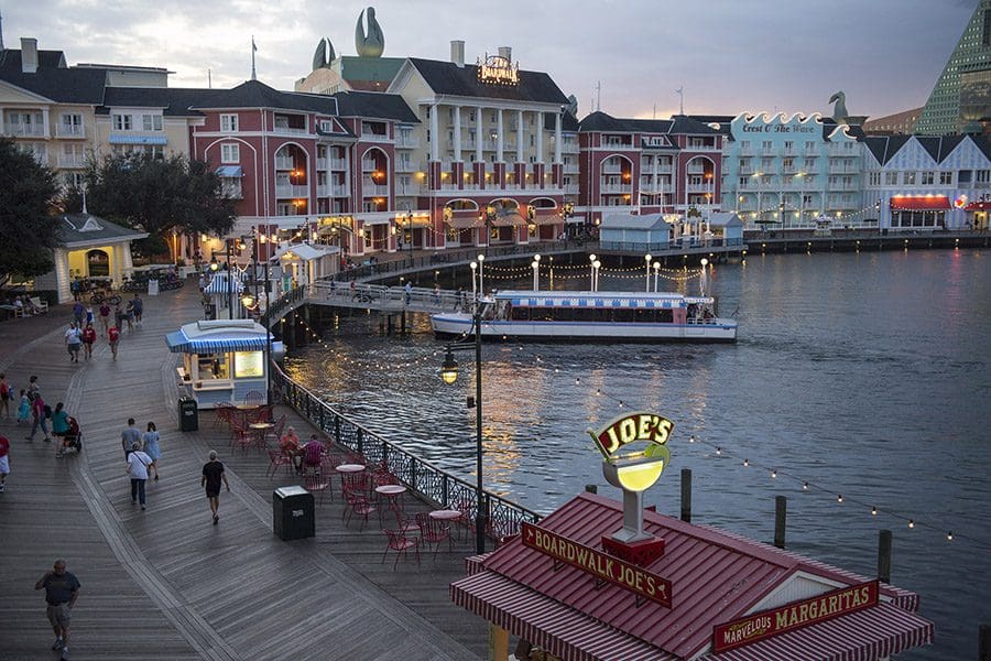 Along the pier at Disney’s Boardwalk Resort, feature a boat on the water and several people wandering the docks.