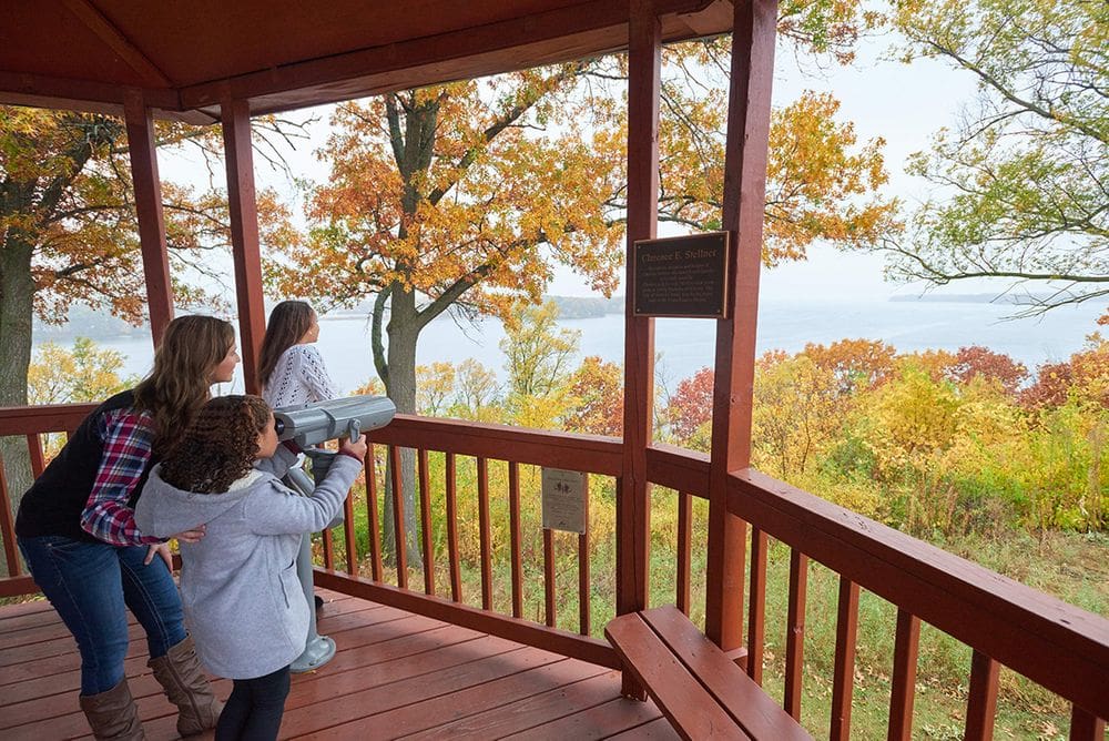 A mom stands near her two children, while one looks through a view finder, and the other stares out onto a beautiful autumn view of Lake Onalaska.