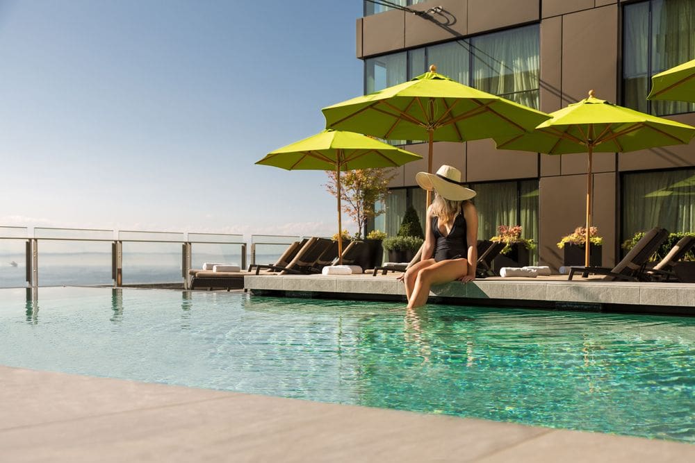 A woman with a large sun hat sits on the edge of the outdoor pool at the Four Seasons Hotel Seattle, with lime green umbrellas and brown pool loungers behind her.
